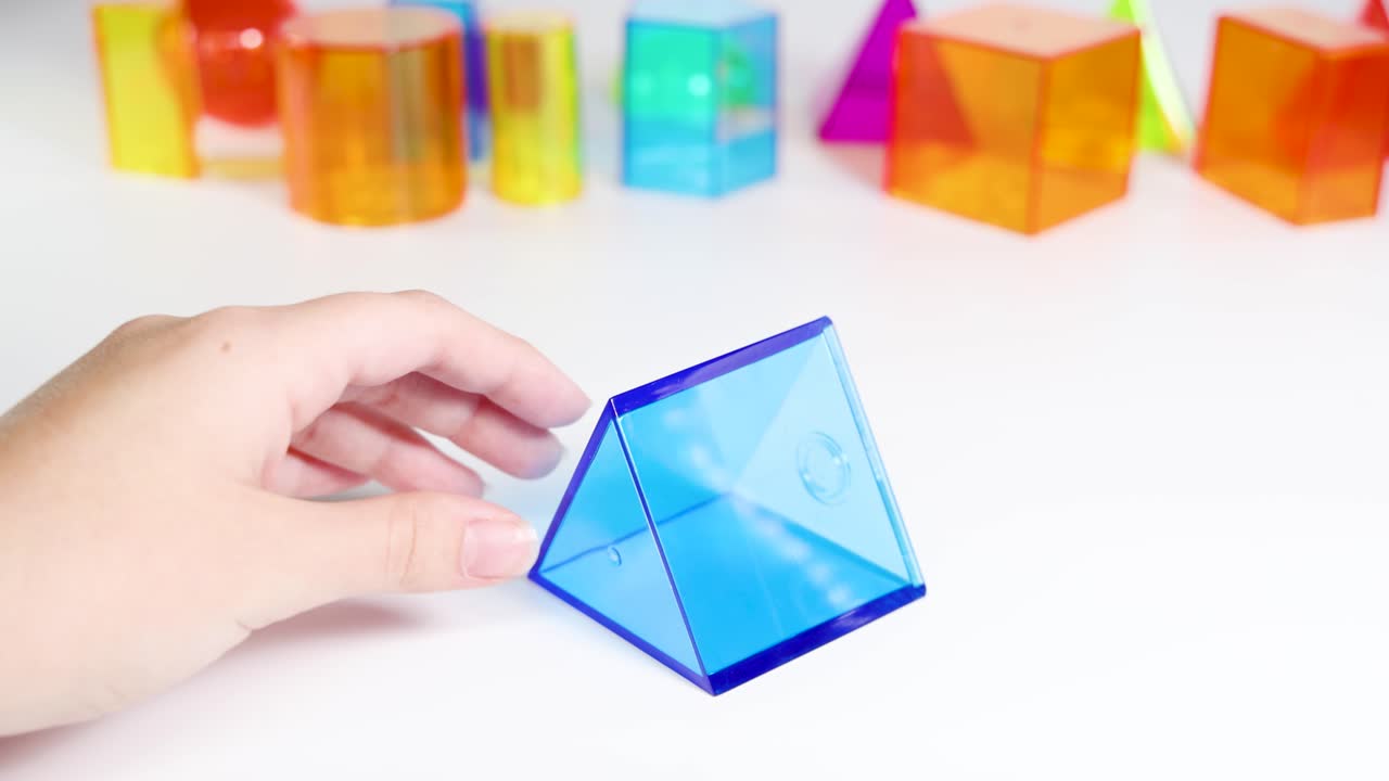 Hands manipulate colorful 3D geometric shapes on a white surface, demonstrating educational engagement with prisms and pyramids