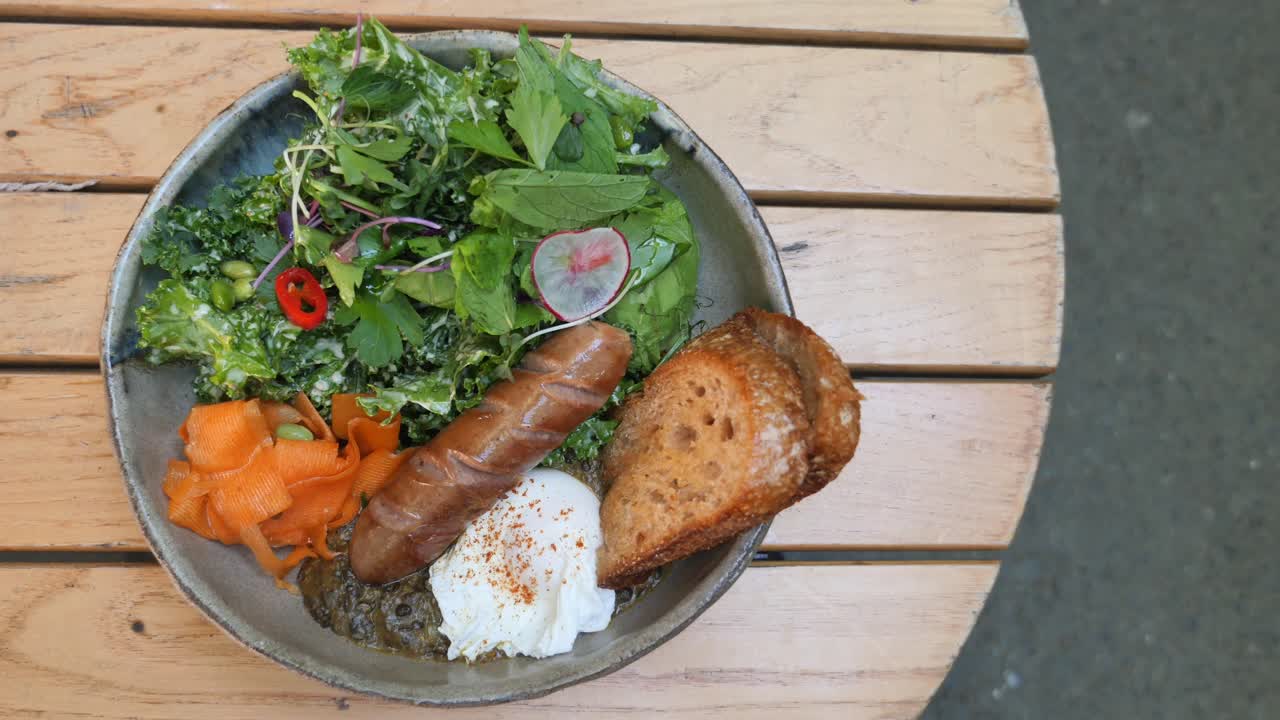 A rustic breakfast or brunch plate featuring sausage, a poached egg, fresh salad, and toast on a wooden table