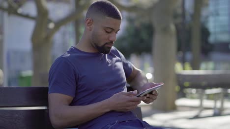 afro-american muscular man on bench in park typing on tablet