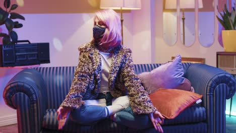 caucasian stylish young woman in fur coat wearing mask sitting on couch in lotus pose practicing yoga and looking at camera in retro room at home