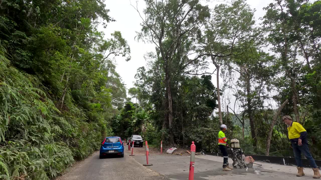 Blue car drives through rainforest roadworks, workers present, overcast daylight, steady forward camera movement