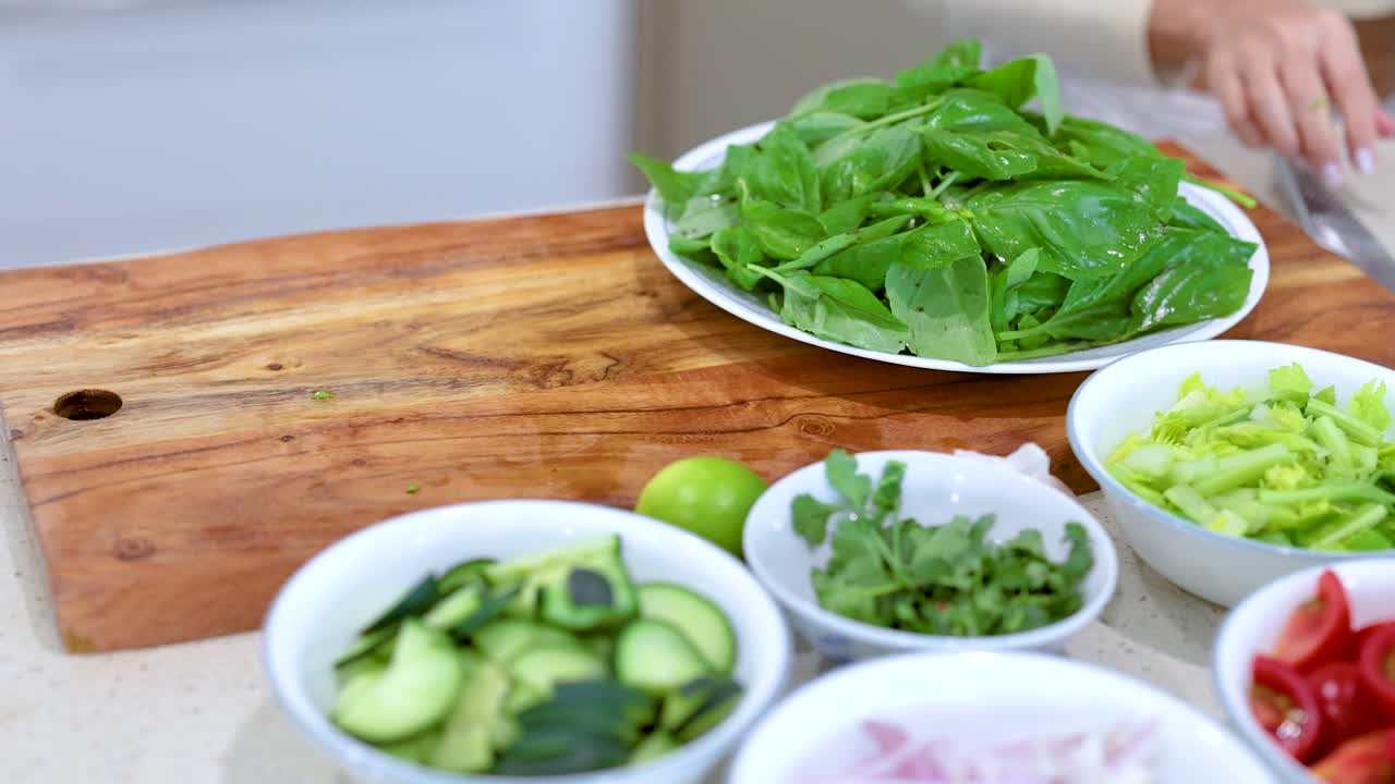 Hands arranging fresh basil and vegetables on a wooden board in a bright kitchen setting