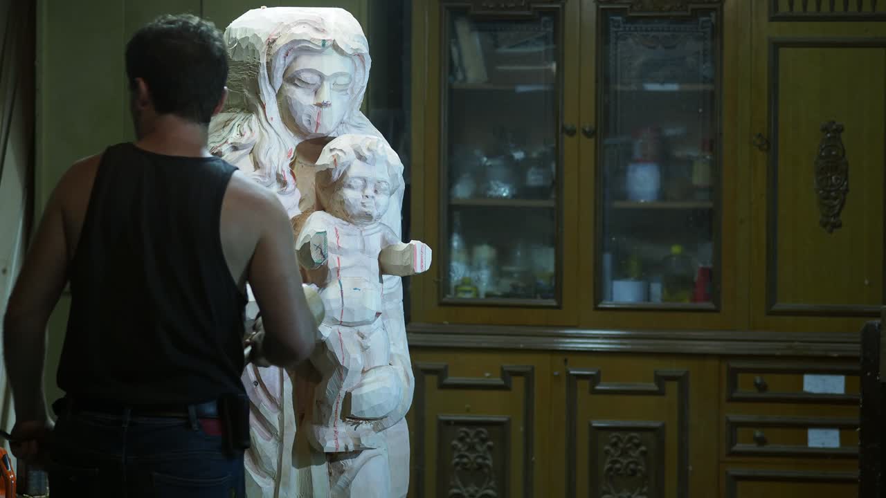 A wood sculptor stands facing a partially carved statue of the Virgin Mary and Child, holding a wooden mallet as he prepares to shape the intricate figure in a traditional indoor workshop setting