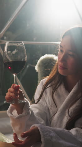 woman holds glass of red wine on glamping terrace. relaxed lady in terry bathrobe enjoys beverage sitting in soft armchair alone by panoramic window at night
