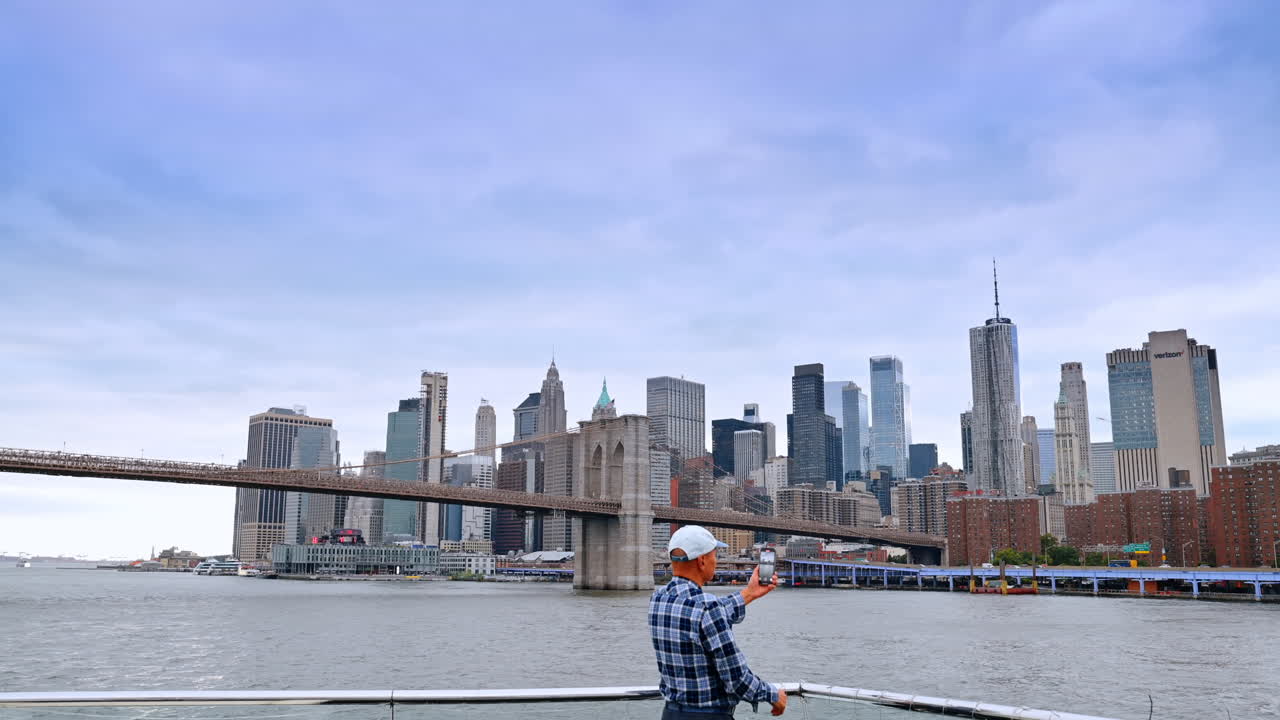 Uomo con cappellino e camicia a quadri che scatta una foto con il suo telefono. Skyline di New York e il Ponte di Brooklyn sullo sfondo