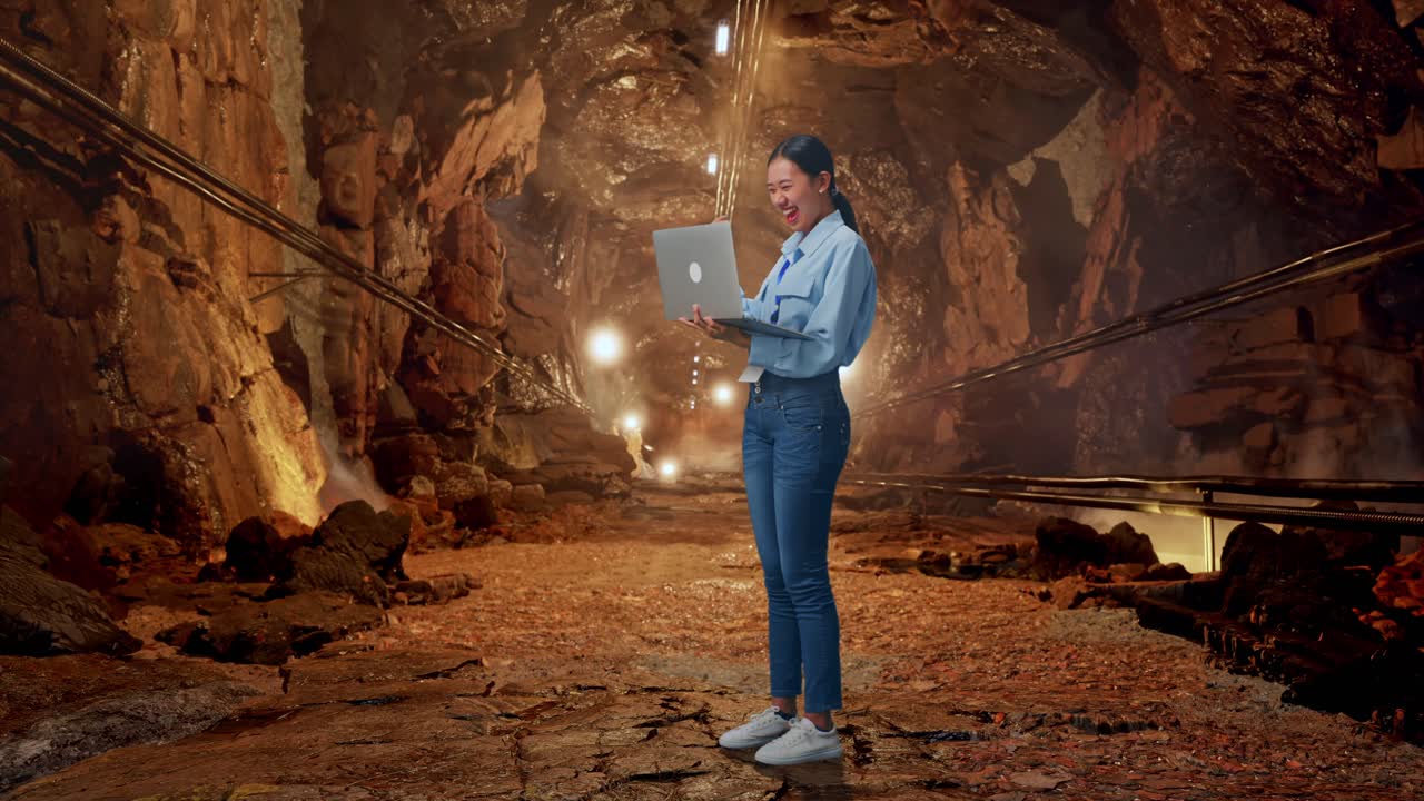 Full Body Side View Of Asian Female Professional Worker With Her Laptop In Underground Mine Tunnel, She Raises Her Fist Up With Screaming Goal After Typed On The Laptop