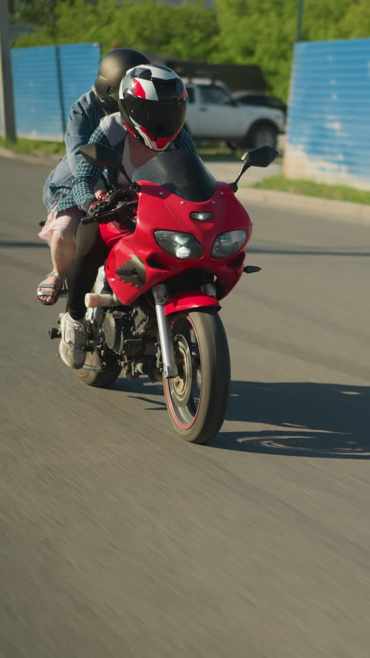 dos hermanas en una bicicleta eléctrica roja viajan a lo largo de una carretera urbana, pasando por un edificio y un coche estacionado visto a través de una puerta de zinc, un coche de color ceniza sigue por detrás, su farol encendido