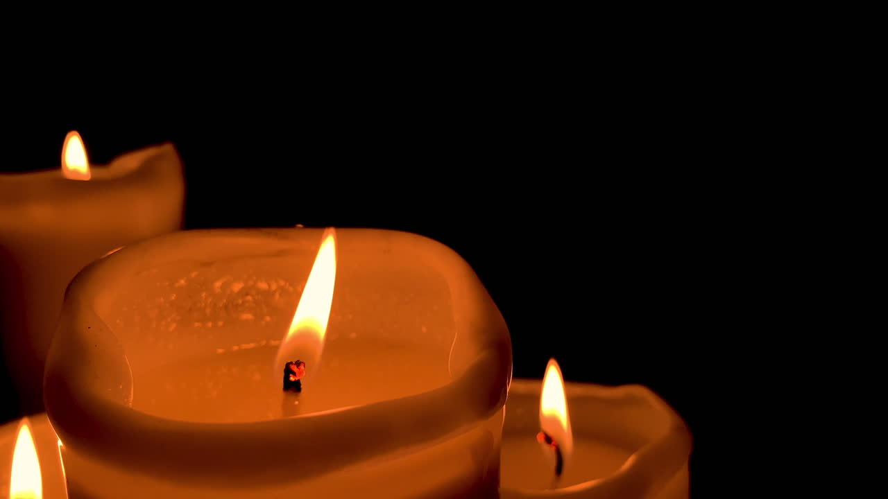 Primo piano su una candela arancione accesa, altre candele sullo sfondo e una parete nera dietro, con la grande fiamma della candela che oscilla al rallentatore