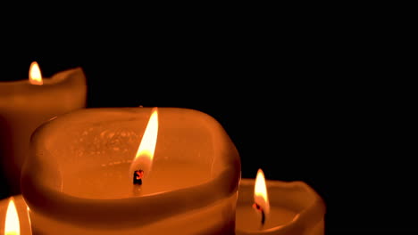 Close-up-at-lit-orange-candle,-other-candles-on-background-and-black-wall-backward,-the-large-candle-flame-swings-in-slow-motion