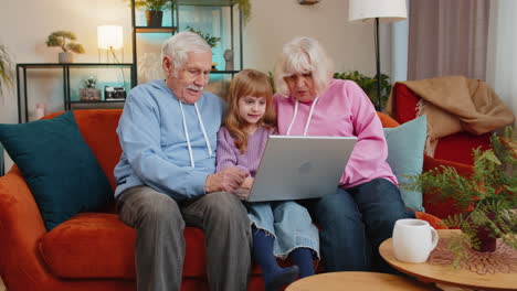 senior couple man and woman with child girl granddaughter spending time at home sofa using laptop
