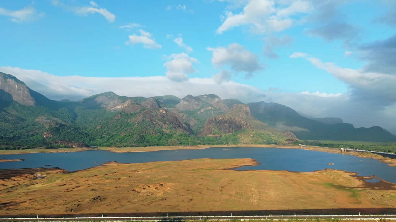 Pemandangan drone udara Waduk Bendungan Aliyar dekat Pollachi, Tamil Nadu, dengan permukaan air rendah yang menyingkap dasar danau kering, pegunungan Western Ghats dan kaki bukit yang menjulang di bawah langit biru berawan di atasnya