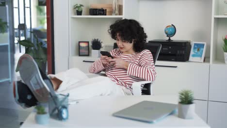 retrato de una hermosa joven de pelo rizado sentada en un escritorio, apoyando los pies en la mesa, jugando un juego atractivo en su teléfono y sonriendo