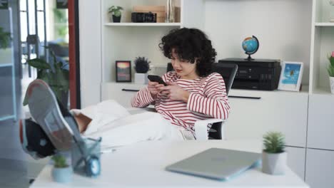 portrait of a beautiful young curly-haired woman sitting at a desk, propping her feet on the table, playing an engaging game on her phone, and smiling