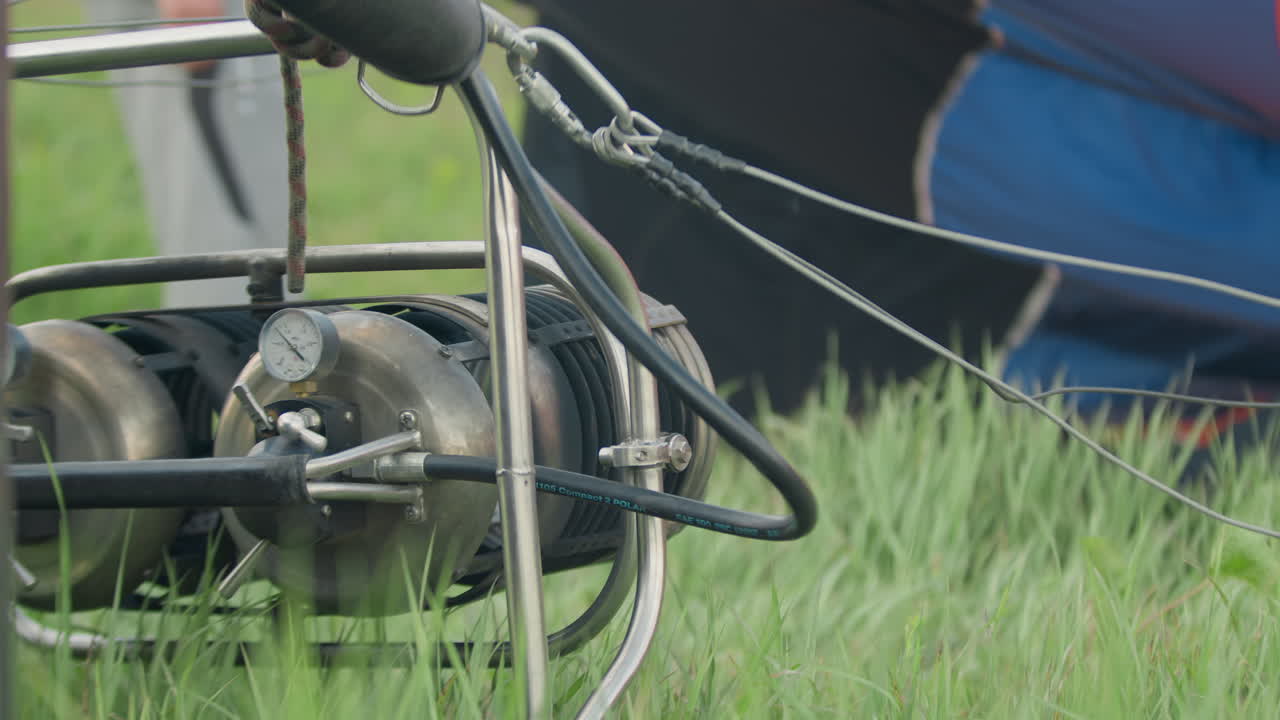 Close up of hot air balloon engine with rotating components on green grass as wires extend outward and blades spin beside swaying vegetation in windy open air during outdoor launch preparation