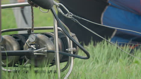 close up of hot air balloon engine with rotating components on green grass as wires extend outward and blades spin beside swaying vegetation in windy open air during outdoor launch preparation