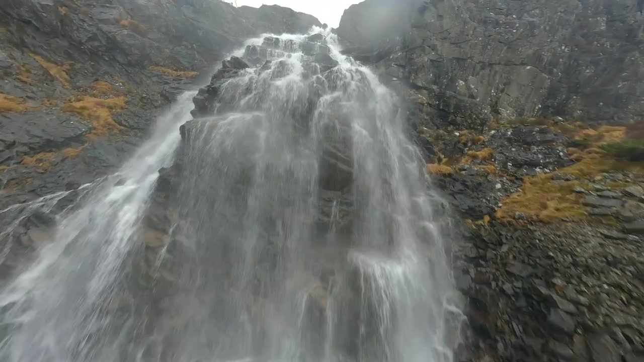 FPV-drone flyr mot en mektig foss i et steinete norsk fjellandskap.