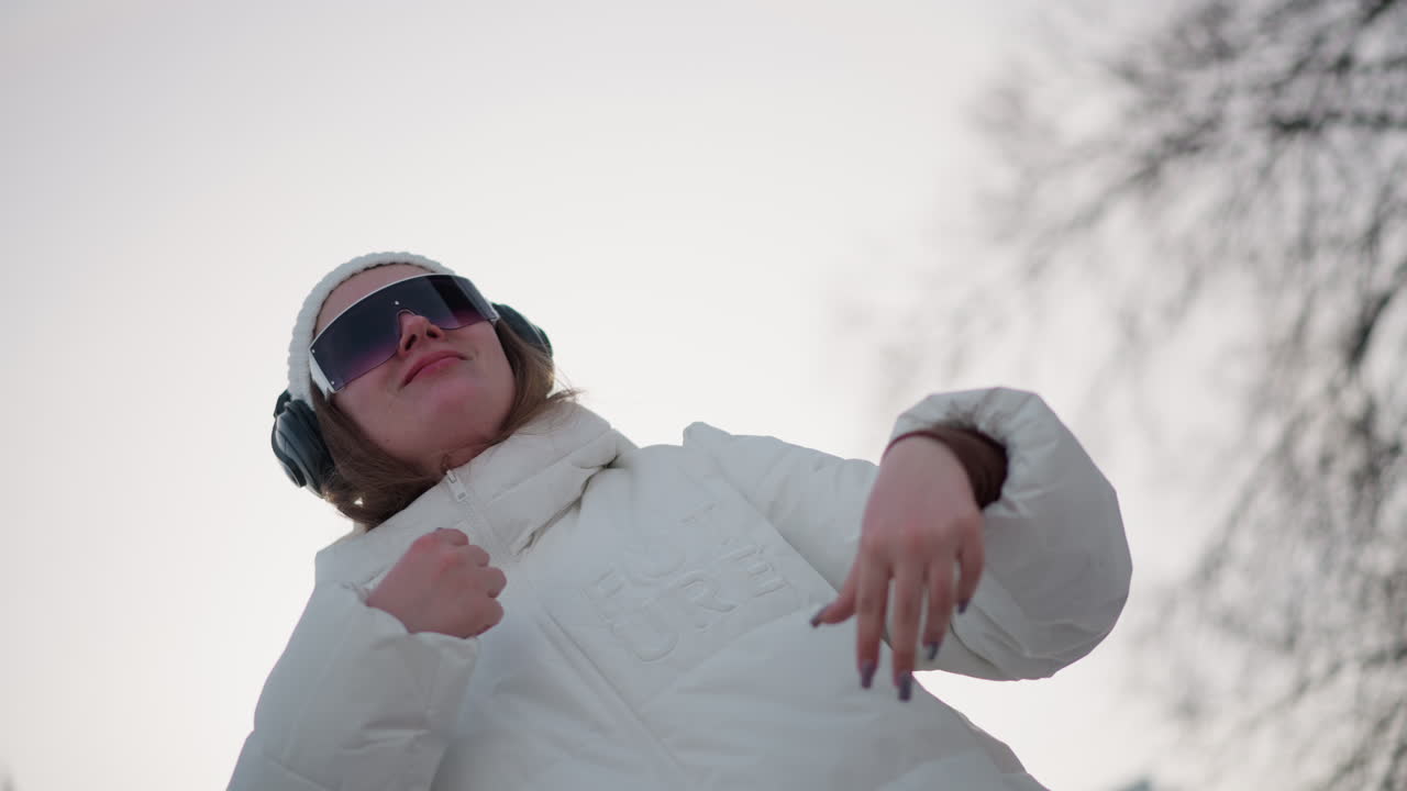Closeup of young artist wearing headphones and beanie, elated and moving with rhythm through fluid choreography on snow dusted urban plaza, hand gestures and joyful smile under pale winter sunlight