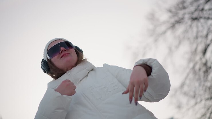 Closeup of young artist wearing headphones and beanie, elated and moving with rhythm through fluid choreography on snow dusted urban plaza, hand gestures and joyful smile under pale winter sunlight