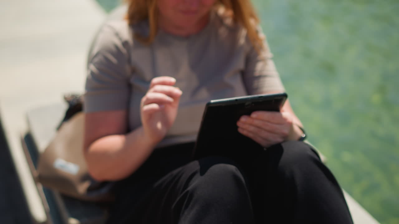 Close up of freelancer smiling while operating tablet under sunlight, gentle warmth on skin, calm outdoor moment by water, relaxed focus and creativity, symbol of modern work blending