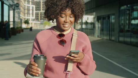 african american woman standing with e-scooter and using smartphone in city