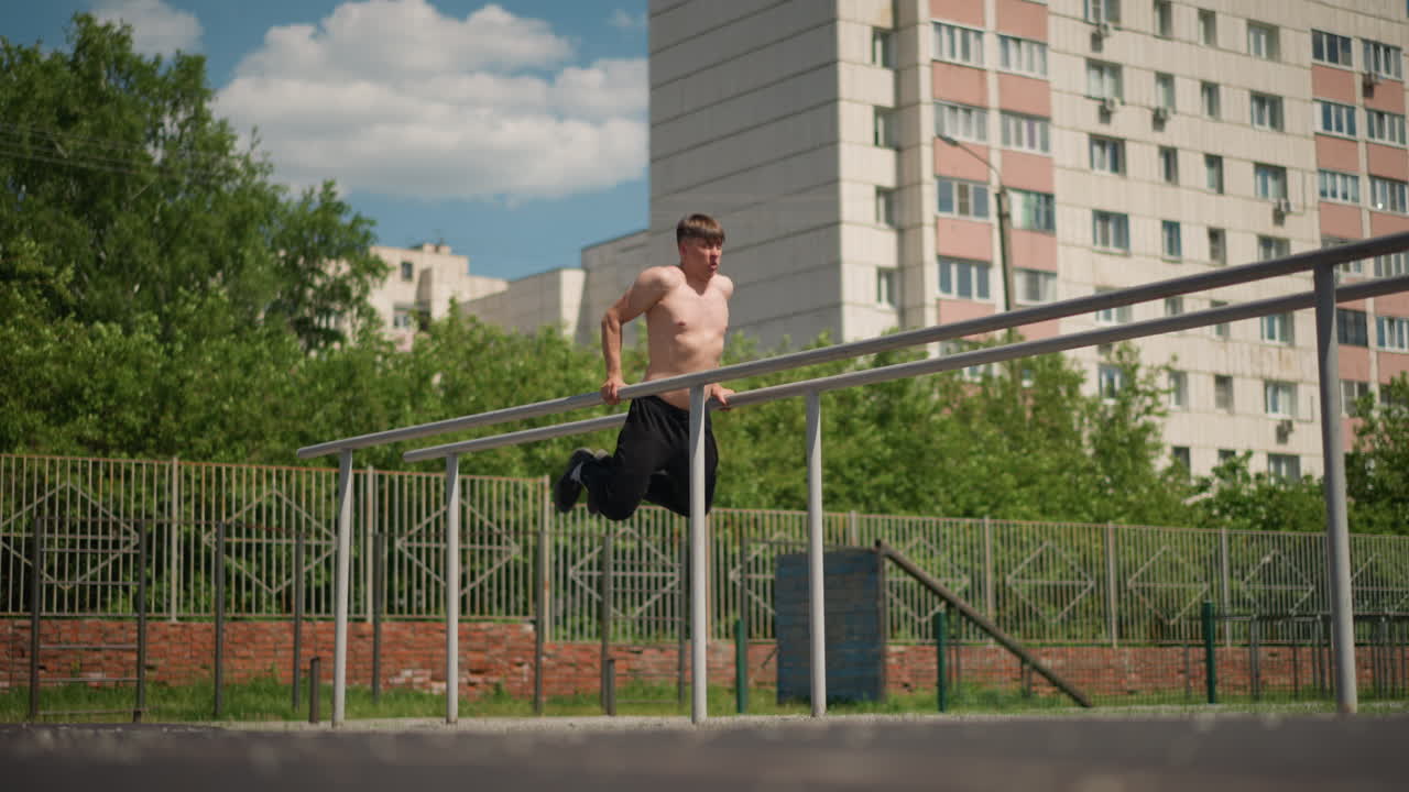 Hombre blanco sin camiseta saltando una valla en un patio, salto y aterrizaje explosivos, obstáculos de hormigón y metal, torres de apartamentos iluminadas por el sol al fondo, momento dinámico de entrenamiento callejero