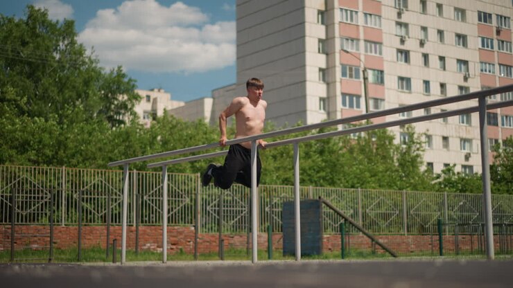 shirtless white man vaulting rail across courtyard fence, explosive leap and landing, concrete and metal obstacles, sunlit apartment towers in background, dynamic street training moment