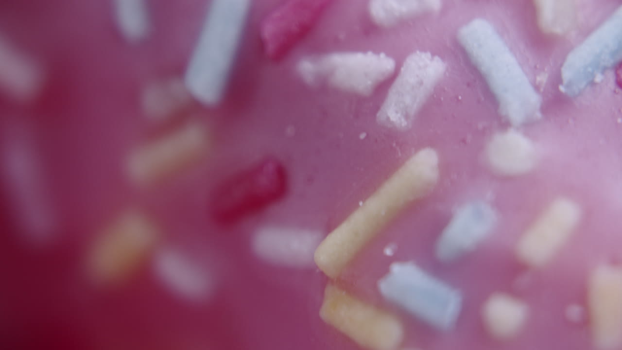 Extreme macro shot of colored sprinkles on top of a pink donut. Donut spins clockwise. Shot with shallow depth of field.