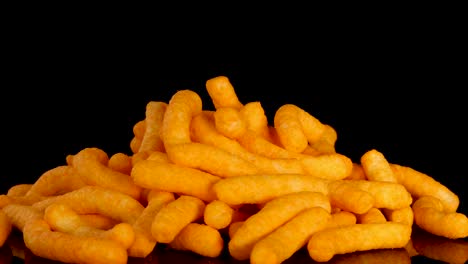 a pile of bright orange cheese puffs snack rotates smoothly against a solid black background, with even studio lighting and a consistent close-up perspective