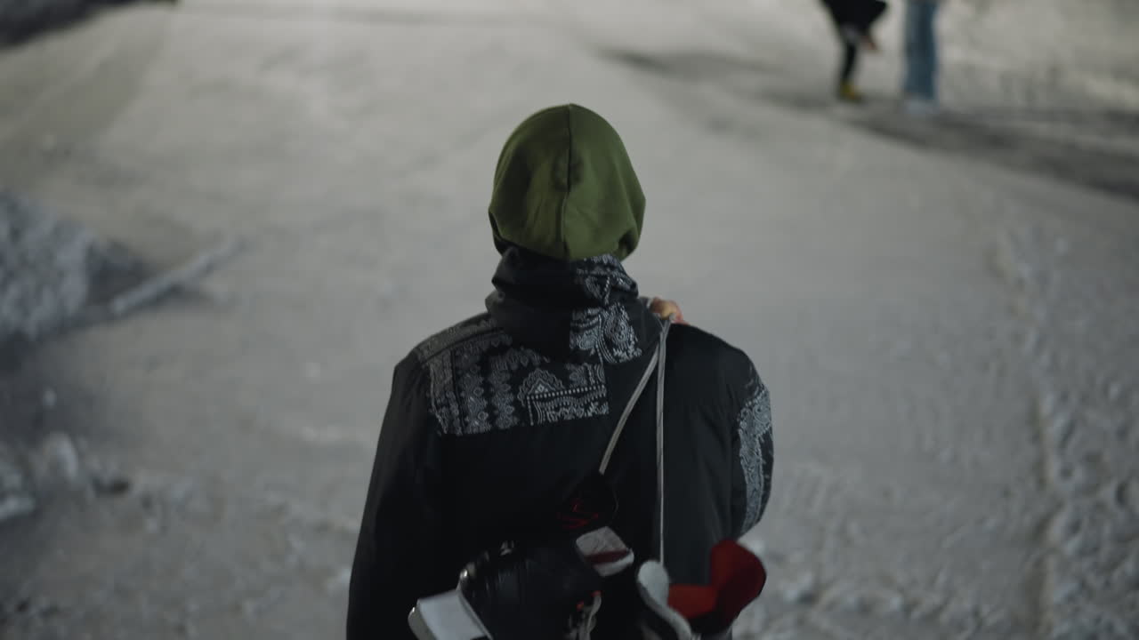 rear view of person in green hood walking on ice rink holding skate boot over shoulder while others stand ahead, one bending to fix skate boot in cold evening environment with snow and icy surface