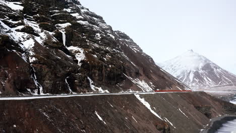 Aerial-View-of-Djúpivogur-Landscape-Iceland,-Car-Driving-Moving-on-the-Edge-of-Rocky-Snowy-Mountain-Road-Trip,-Búlandstindur-Basalt-High-Mountain-Covered-by-Snow-in-Horizon