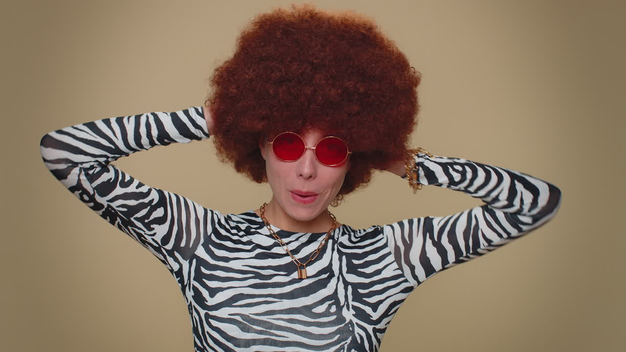 alegre hermosa joven con peluca marrón exuberante en gafas de sol rojas sonriendo, mirando a la cámara