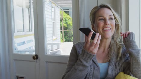 Happy-caucasian-mature-woman-siting-in-living-room-and-using-smartphone