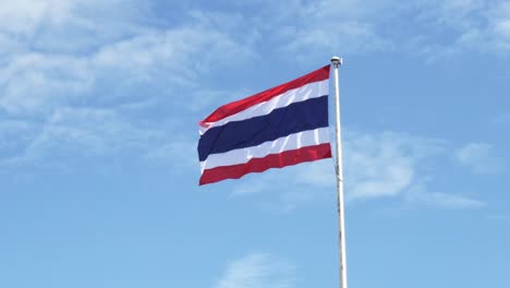 Thailand-national-flag-hoisted-on-a-white-tall-steel-pole-blown-by-the-wind-creating-an-animated-wave-on-a-backdrop-of-a-clear-blue-sky-and-white-clouds