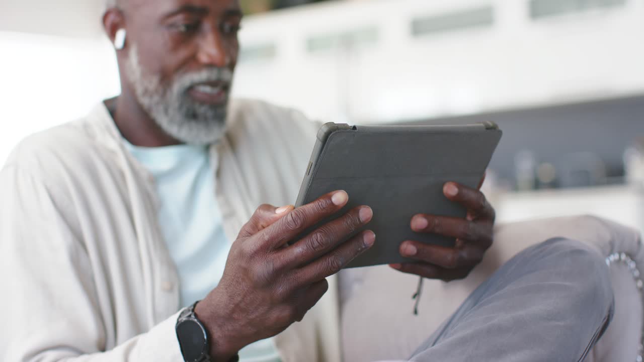 Starší muž afrického původu drží tablet na pohovce, upravuje úchop, ťuká do něj a usmívá se na obsah na obrazovce.