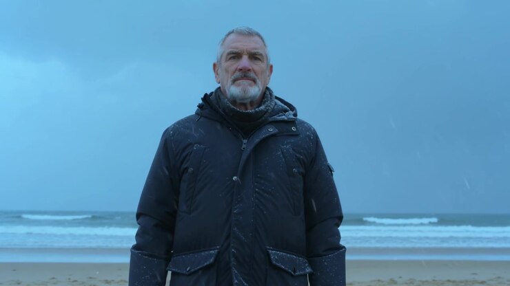En eldre mann strammer skjerfet, vender mot kamera på en sandstrand mens sjøsprøyt pisker mot parkasen.