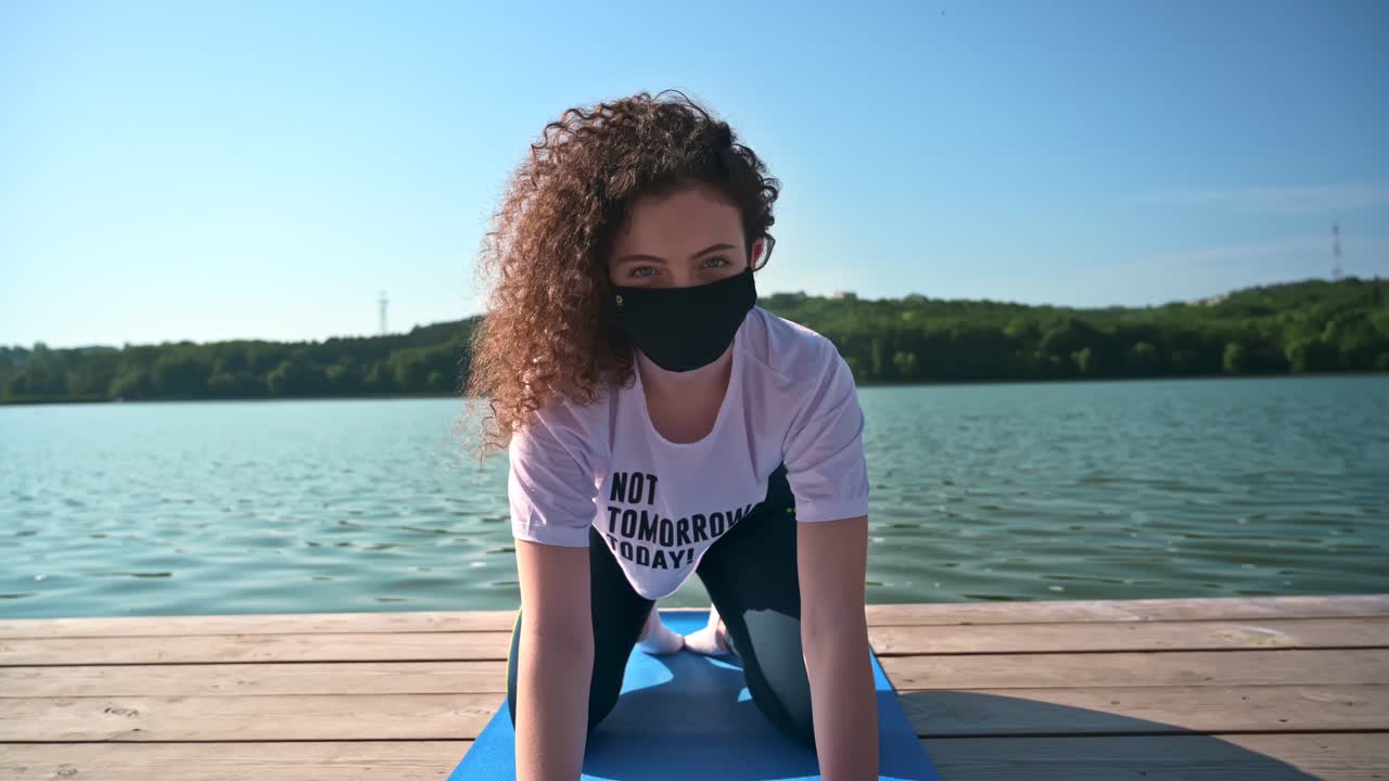 Young curly woman practicing yoga near the lake while wearing black protective mask. Sunny day. Meditating in child's pose on the yoga mat. Corona Virus idea. Chisinau, Moldova