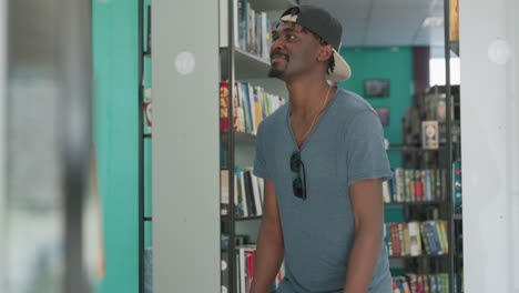 Happy college student wearing backward cap and casual t shirt dancing playfully near library bookshelves, surrounded by colorful books in bright reading space with teal walls and relaxed atmosphere