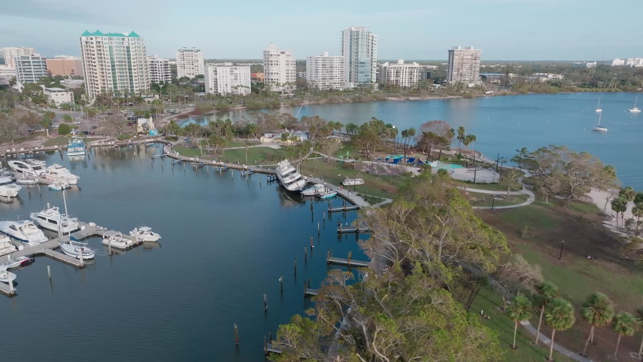 Aerial close-up of grounded yacht in Bayfront Park, pulling back to reveal Sarasota skyline, Marina Jack, and Sarasota Bay. Pelican flies past camera as it orbits anchored boats post-Hurricane Milton.
