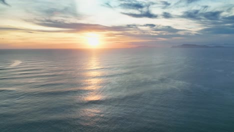 Panning-drone-shot-of-a-beautiful-golden-sunset-reflecting-on-gentle-open-ocean-waves,-with-distant-tropical-island-on-the-horizon