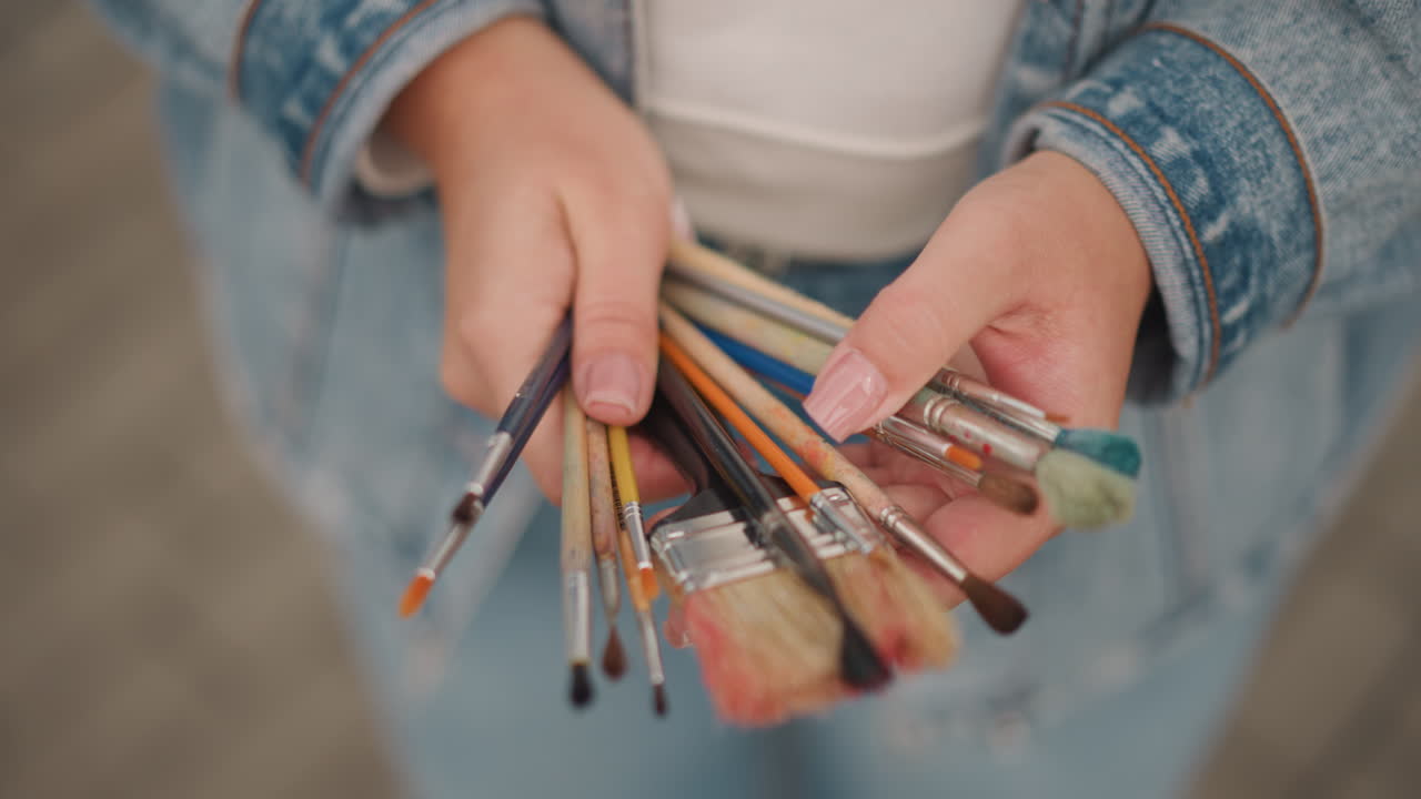 Kunstnerhænder holder pensler tæt på en denimjakke, der viser børster og blandede farver. Tankefuld forberedelse af pensler til atelier-session, sortering af størrelser, test af strøg, blanding af lille palet.