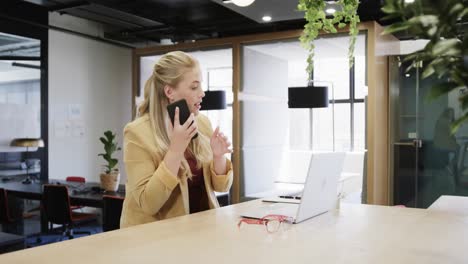 Happy-plus-size-caucasian-casual-businesswoman-using-laptop-and-talking-on-smartphone-at-desk