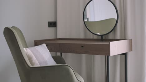 a minimalist dressing area featuring a sleek wooden console table with a round mirror, complemented by a plush olive green chair and a soft pillow, creating a tranquil corner in a modern bedroom.