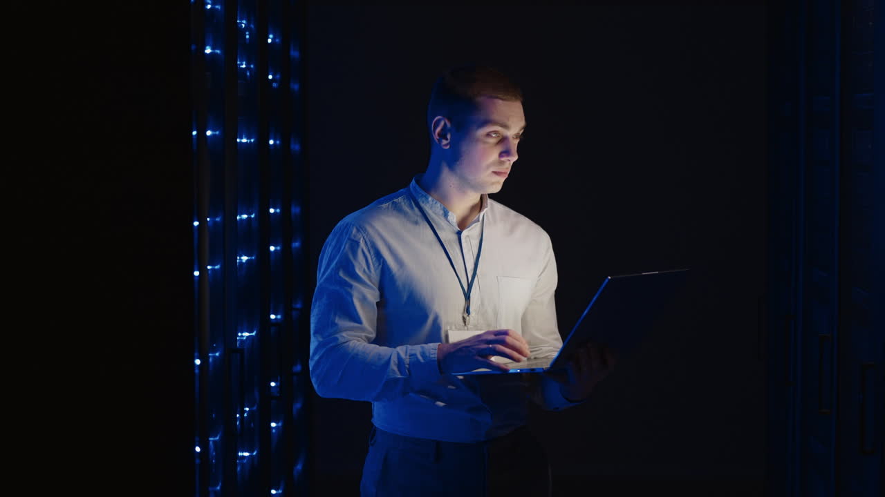 Young man holds device in hand and looks at screen inspects equipment or hardware rack. Male programmer working with laptop and supporting service while standing in data center spbas.