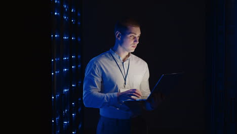 young man holds device in hand and looks at screen inspects equipment or hardware rack. male programmer working with laptop and supporting service while standing in data center spbas.