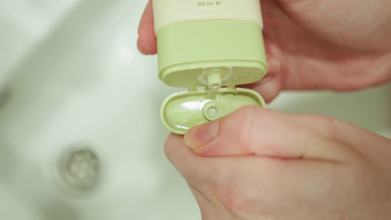 Close-up of hands operating pale green flip-top dispenser above white ceramic sink, visible action shows bottle tilted and thumb lifting lid or pressing to prepare dispensing while clear liquid