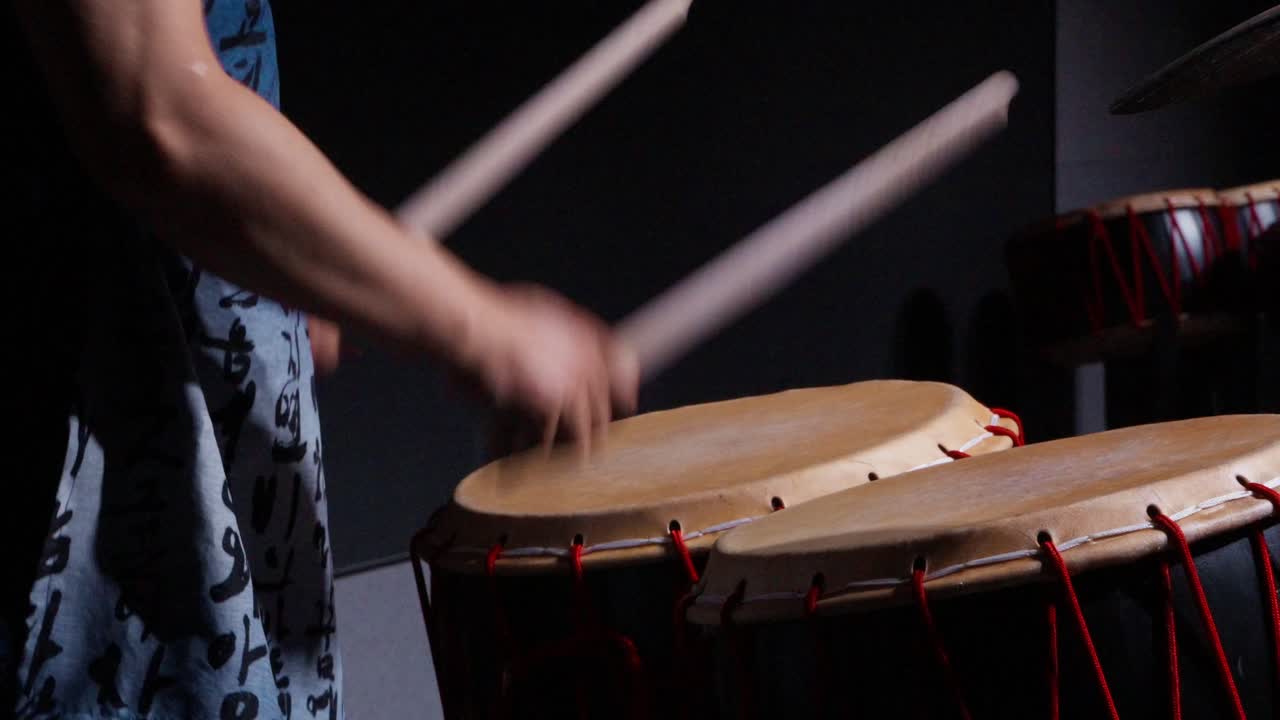 Close-up of hands striking a traditional Korean buk drum with wooden sticks, showing drumhead and red lacing detail. Close-up shot