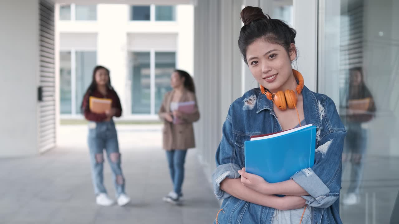 Portrait of an Asian college student holding folders and looking at the camera while standing at the hallway. Education concept.