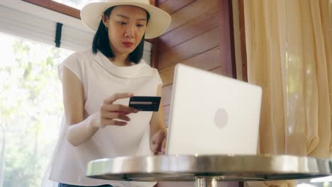 beautiful asian woman sitting in front of the window she holding credit card and using laptop with instant easy mobile payments making purchase in online store. e-banking app.
