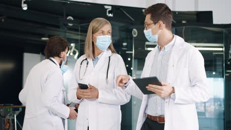 Male-and-female-doctors-in-medical-masks-at-work-walking-and-discussing-healthcare-issues-using-smartphone-and-tablet-device-in-hospital