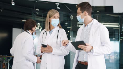 male and female doctors in medical masks at work walking and discussing healthcare issues using smartphone and tablet device in hospital